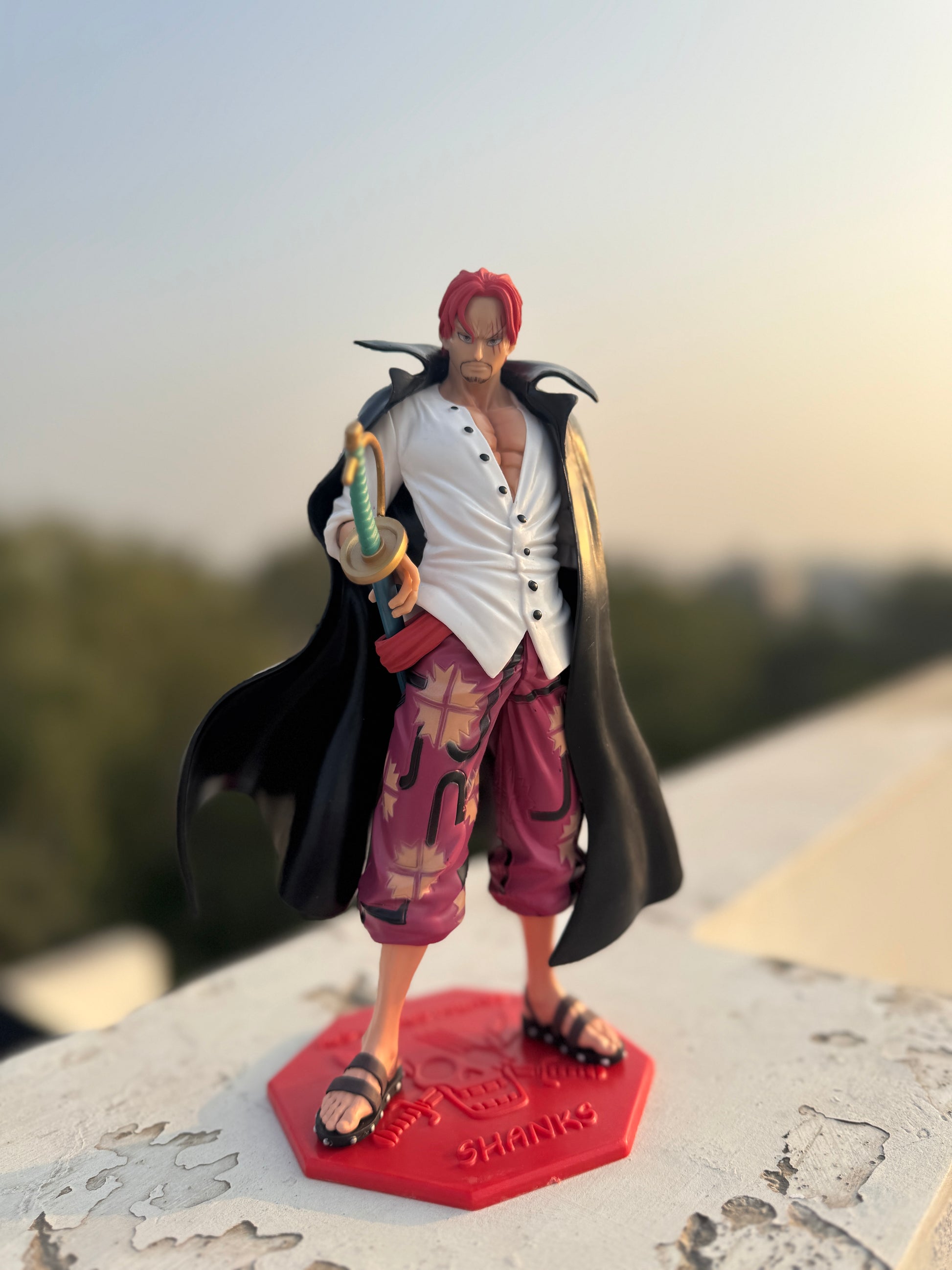 Action figure of a character with red hair and a black cloak on a red base with 'Shanks' branding, outdoors.