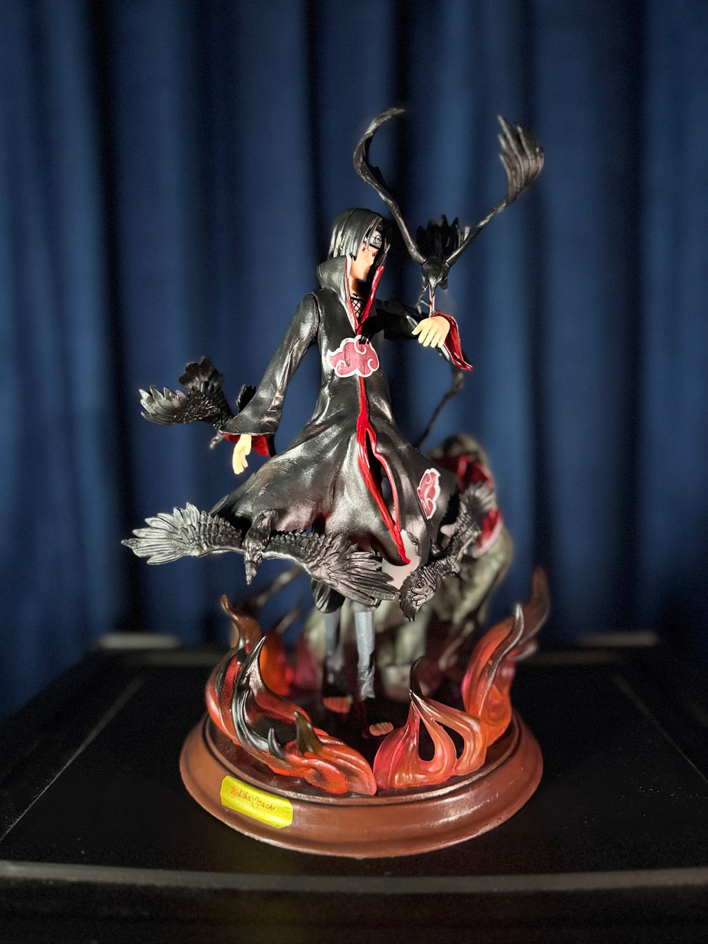 Detailed figurine of a character with a dark outfit and red accents on a wooden base against a blue curtain background.
